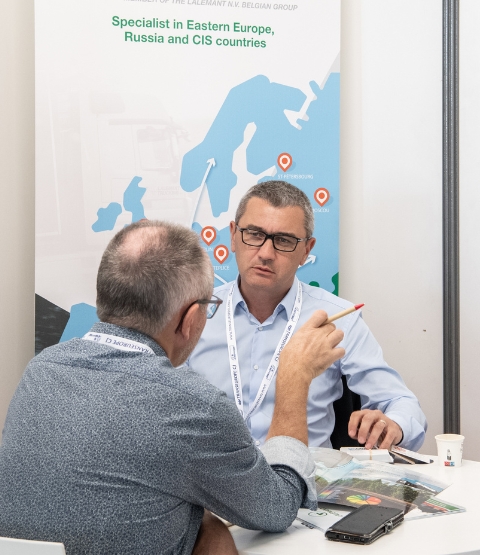 Conversation between two men during a one-to-one meeting at Top Transport Europe, with documents and a map on the wall.